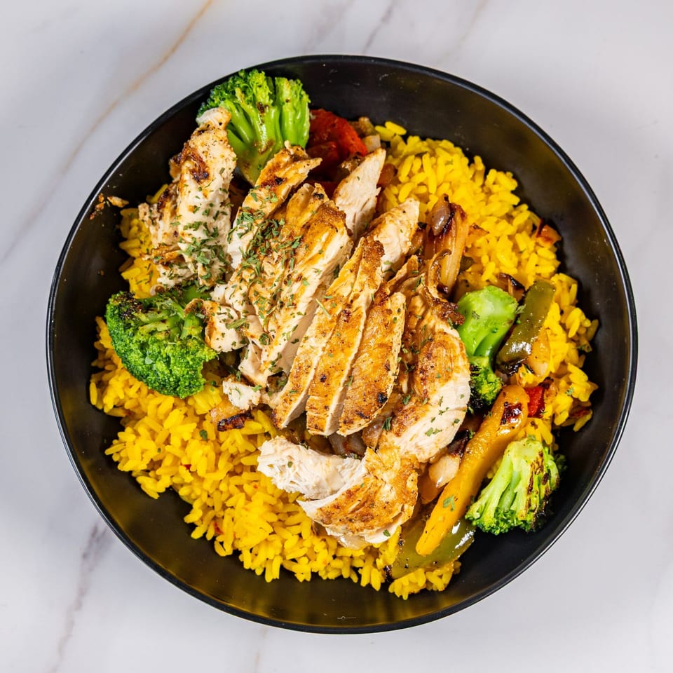 Grilled chicken slices, yellow rice, and roasted vegetables in a black bowl on a marble surface.