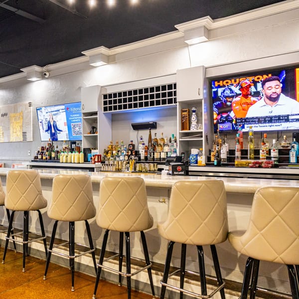 A well-stocked bar with multiple liquor bottles, bar stools, and two TVs showing sports and an ad.