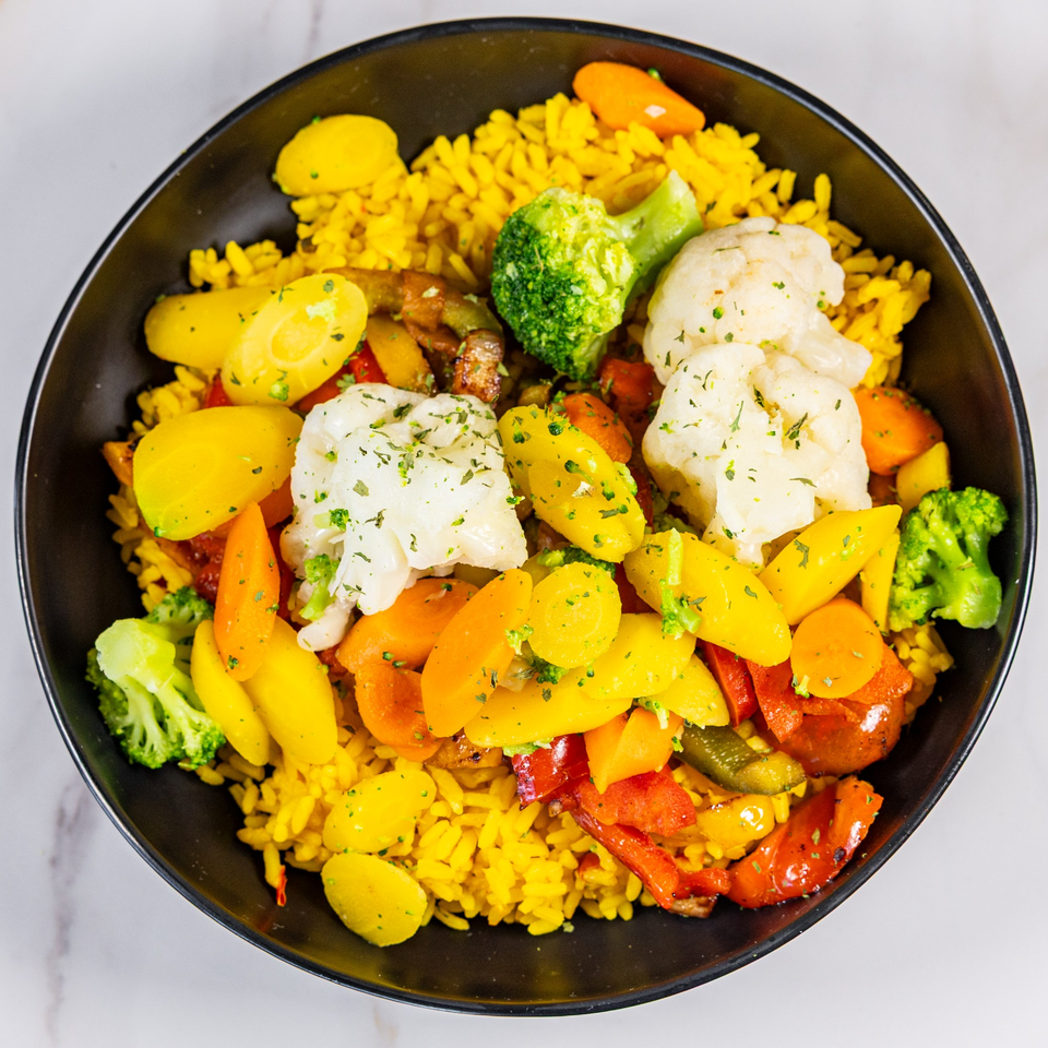 A black bowl filled with yellow rice, roasted vegetables, and herbs on a white marble surface.